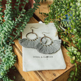 A pair of gray tassel earrings displayed on a white card with the brand name 'Steel & Clover' surrounded by green plants.