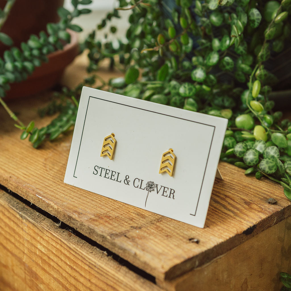 A pair of gold-plated arrow stud earrings displayed on a card with the brand name Steel & Clover, placed on a wooden surface surrounded by greenery.