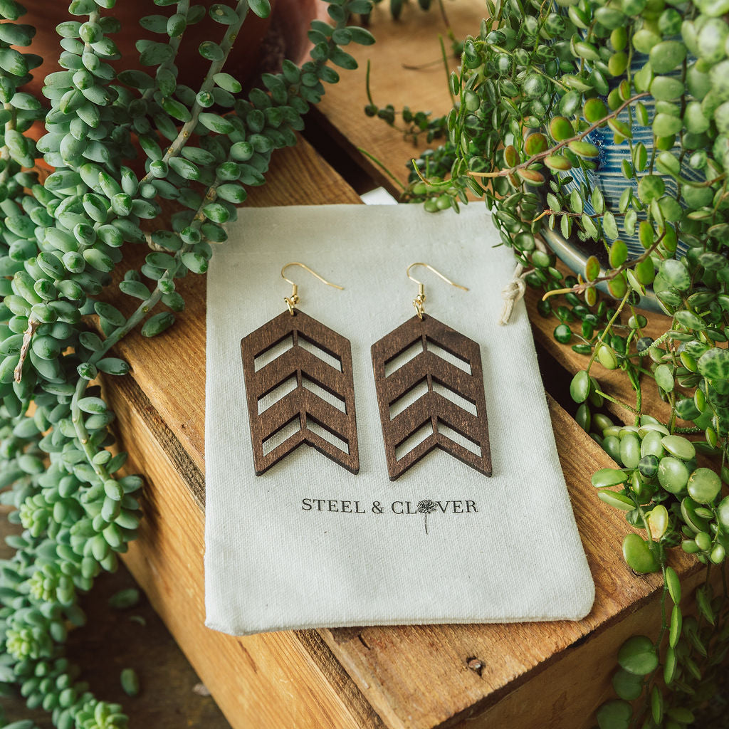 A pair of wooden, geometric arrow-shaped earrings displayed on a white fabric with the brand name 'Steel & Clover' visible, set against a wooden background with greenery in the backdrop.