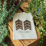 A pair of wooden, geometric arrow-shaped earrings displayed on a white fabric with the brand name 'Steel & Clover' visible, set against a wooden background with greenery in the backdrop.