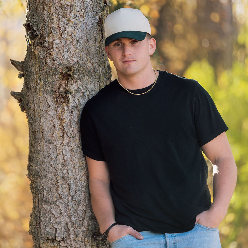 Man wearing a black t-shirt and white cap leaning against a tree in a natural setting