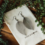 A pair of copper electroplated leaf earrings, with lacey veins, displayed on a white pouch bearing the brand name 'Steel & Clover'. Surrounding the pouch are green leaves.