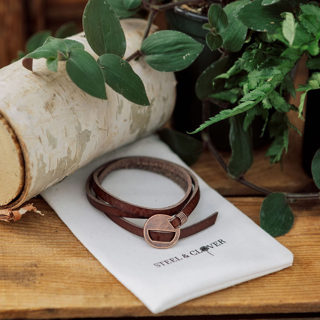A orange and brown leather wrap bracelet with a antique brass grommet, displayed on a white card with the brand name 'Steel & Clover'.