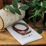 A orange and brown leather wrap bracelet with a antique brass grommet, displayed on a white card with the brand name 'Steel & Clover'.