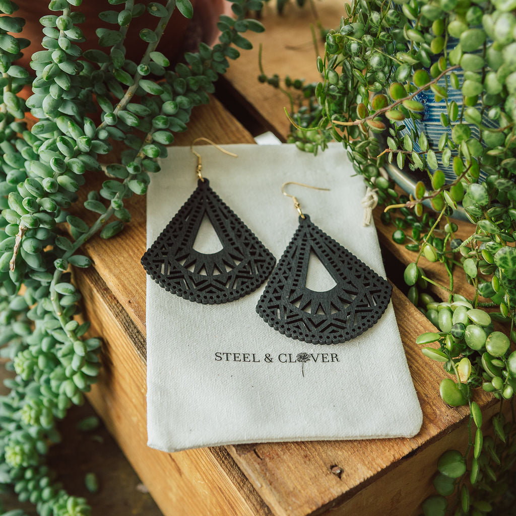 A pair of black wooden earrings with geometric patterns displayed on a beige fabric with the brand name 'Steel & Clover' visible.