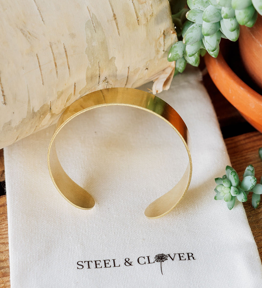 A gold-colored brass cuff bracelet displayed on a textured white background with a brand tag reading 'Steel & Clover'.