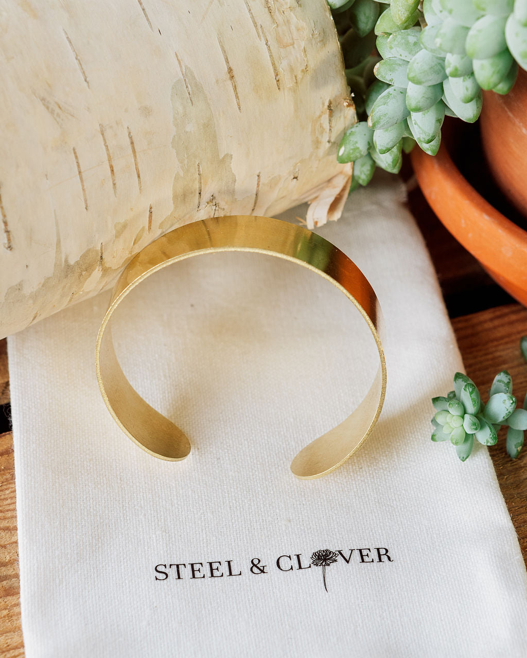 A gold-colored brass cuff bracelet displayed on a textured white background with a brand tag reading 'Steel & Clover'.