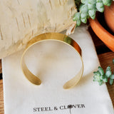 A gold-colored brass cuff bracelet displayed on a textured white background with a brand tag reading 'Steel & Clover'.