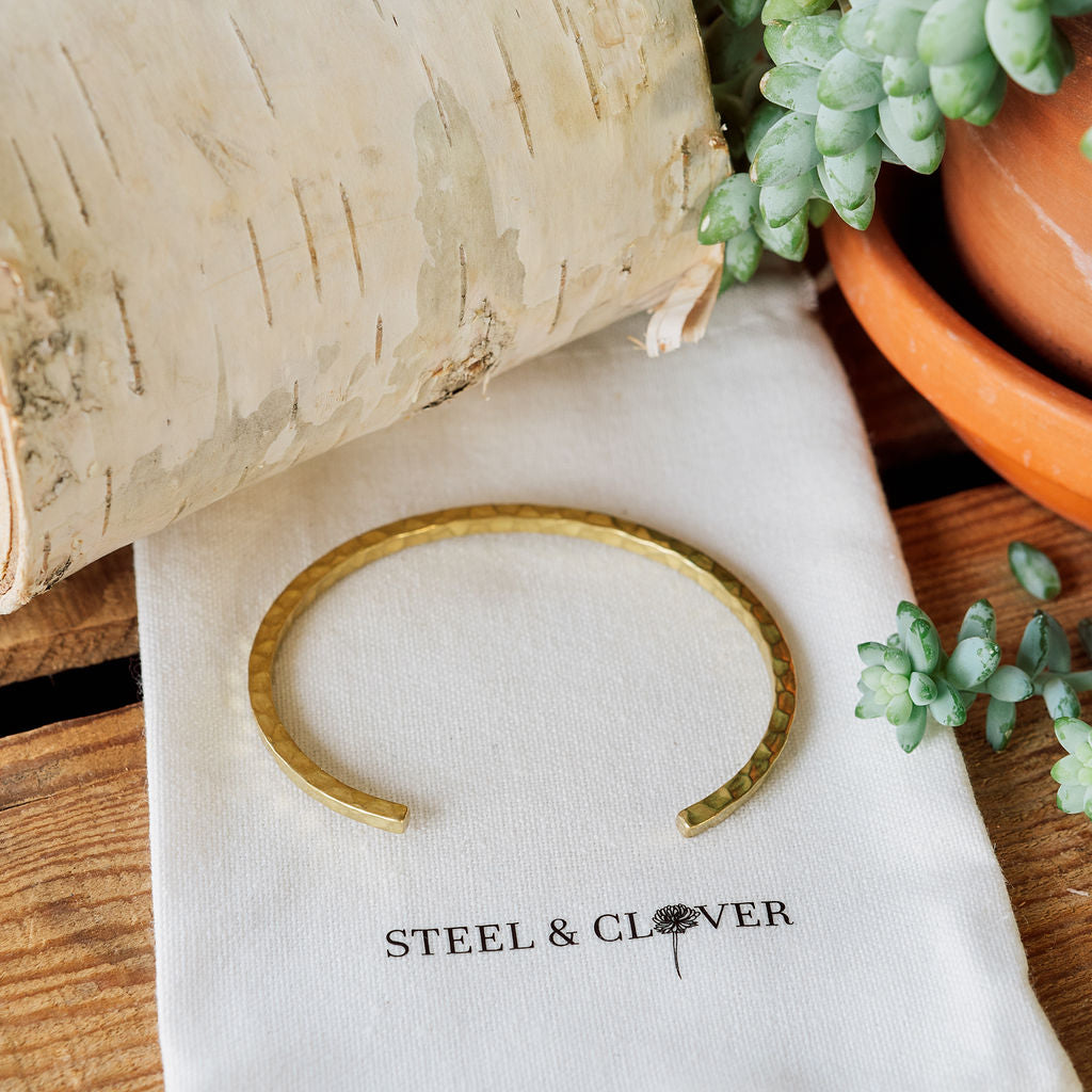 Gold bracelet on a white cloth with 'Steel & Clover' branding, surrounded by plants.