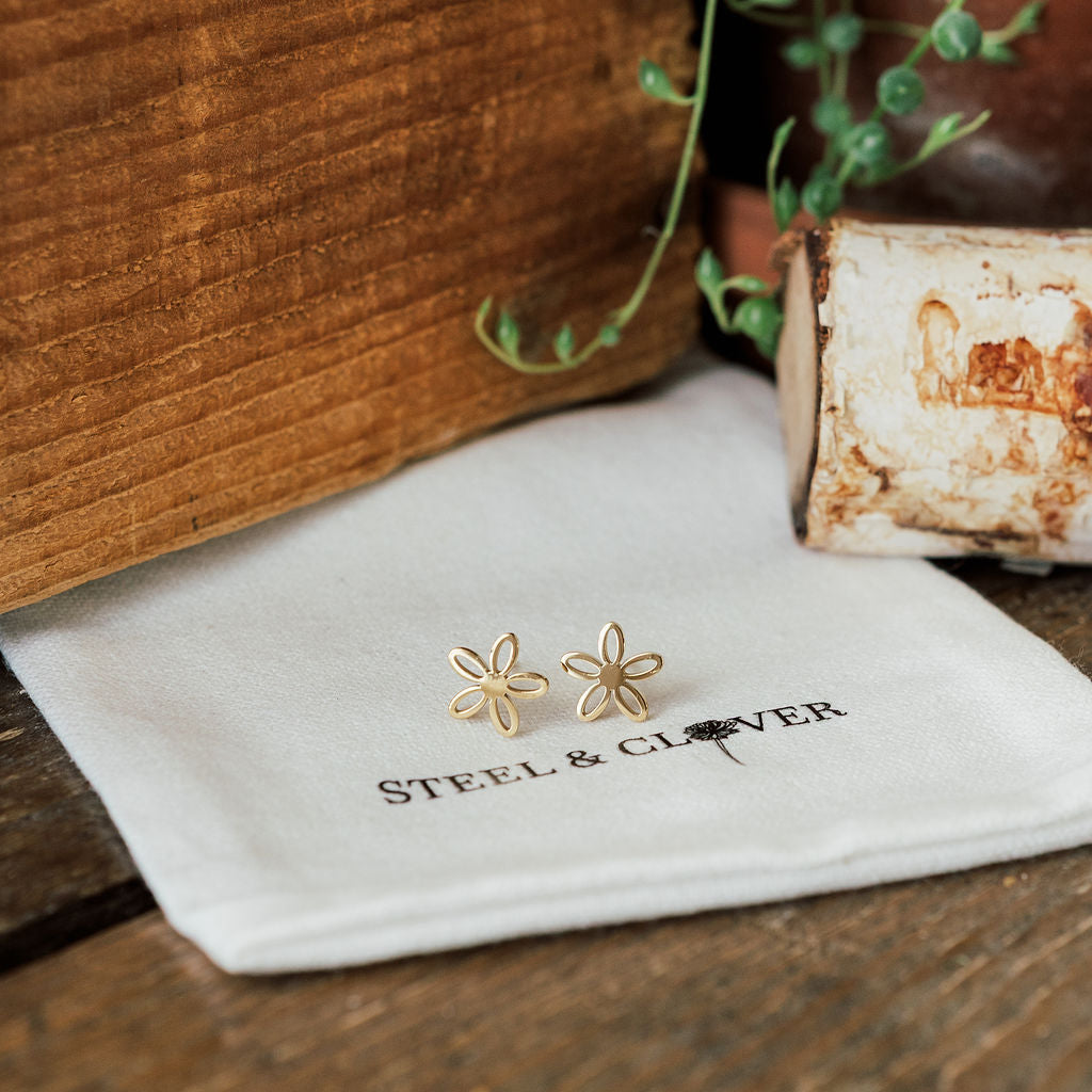 Gold flower-shaped earrings on a 'Steel & Clover' branded cloth with a wooden background.