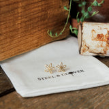 Gold flower-shaped earrings on a 'Steel & Clover' branded cloth with a wooden background.