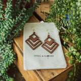 A pair of wooden, geometric earrings displayed on a white card with the brand name 'Steel & Clover' surrounded by greenery.