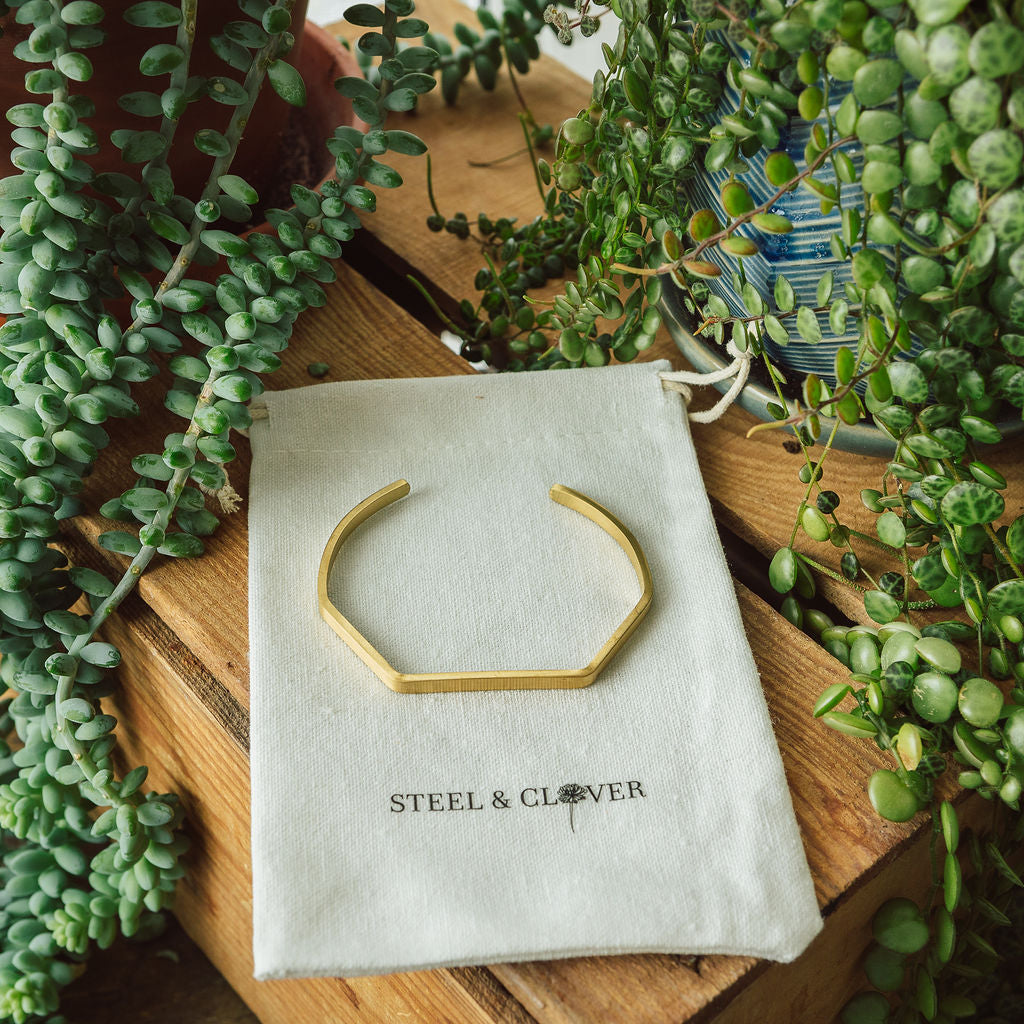 A gold-colored geometric bracelet displayed on a white card with the brand name 'Steel & Clover', surrounded by green plants.