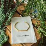 A gold-colored geometric bracelet displayed on a white card with the brand name 'Steel & Clover', surrounded by green plants.