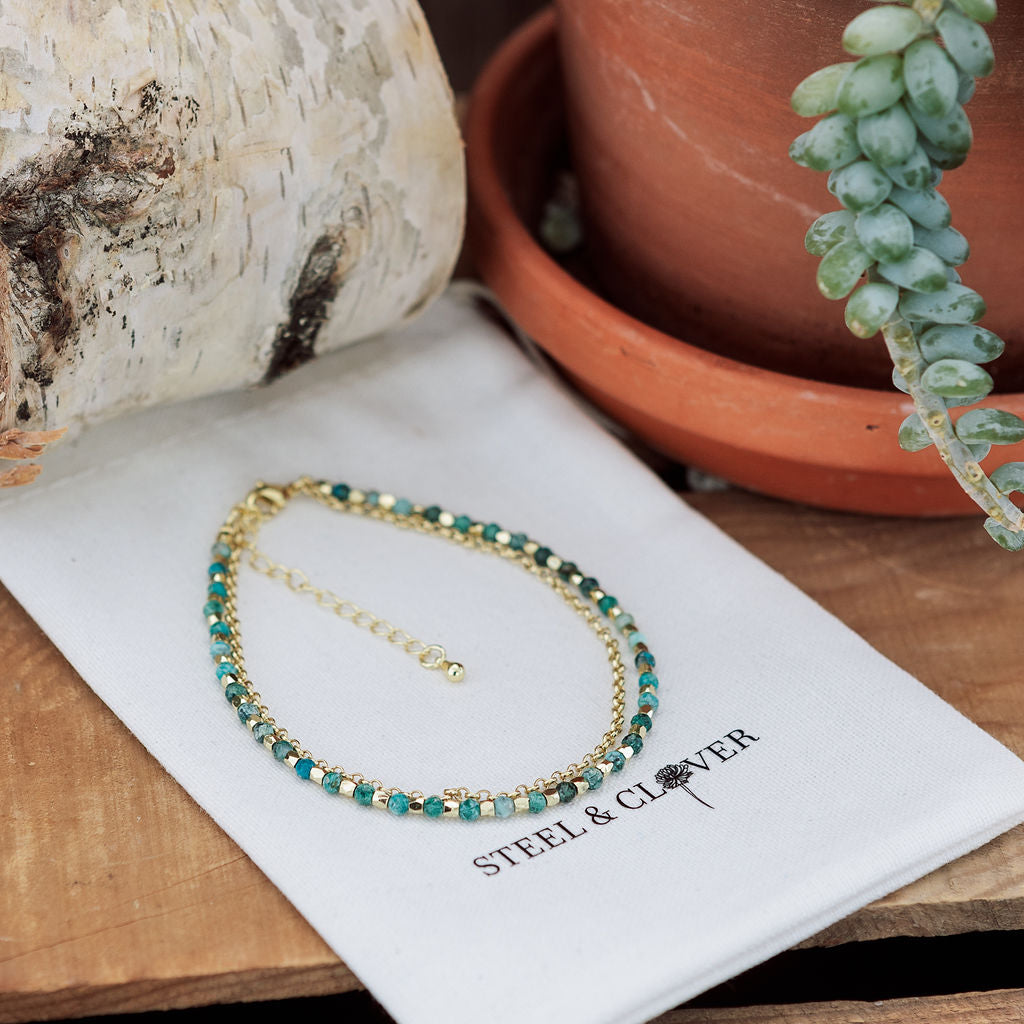 A gold-toned chain anklet with green beaded details laid out on a white surface, with a plant and a birch bark in the background.