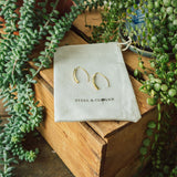 A pair of minimalist hook earrings made from stainless steel, displayed on a beige fabric pouch with the brand name 'Steel & Clover' embroidered on it, placed among green plants.