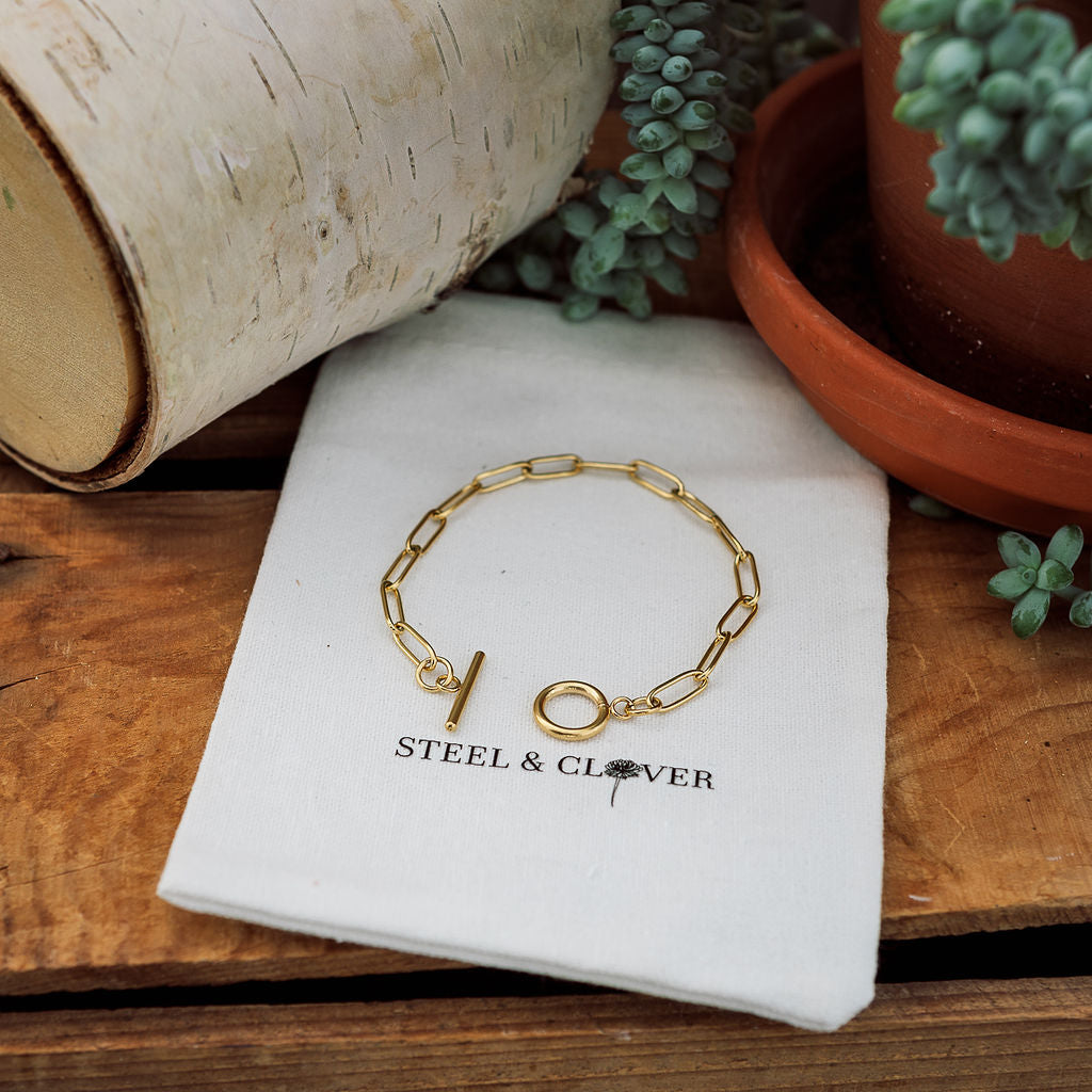 A gold-colored paperclip chain bracelet displayed on a white fabric surface, with a branded Steel & Clover tag visible.