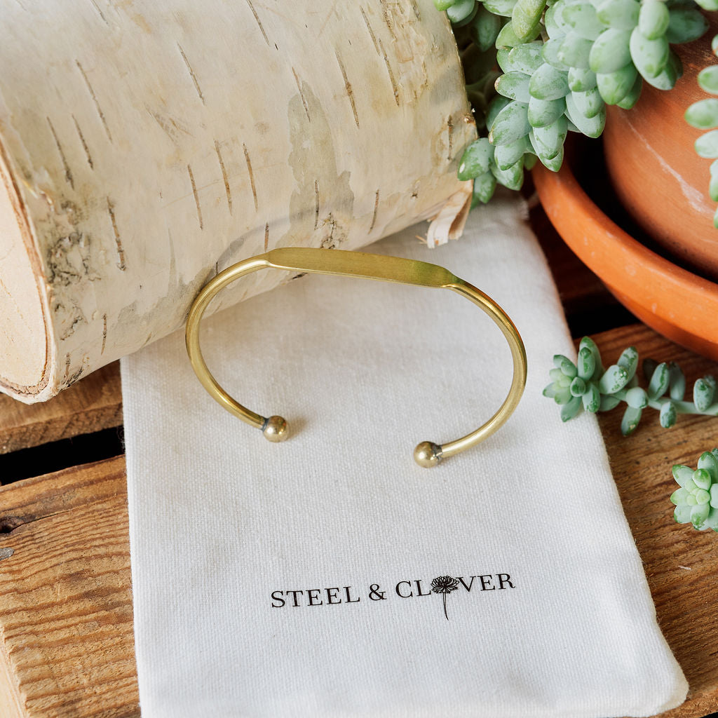 Gold bracelet on a 'Steel & Clover' branded pouch with plants in the background