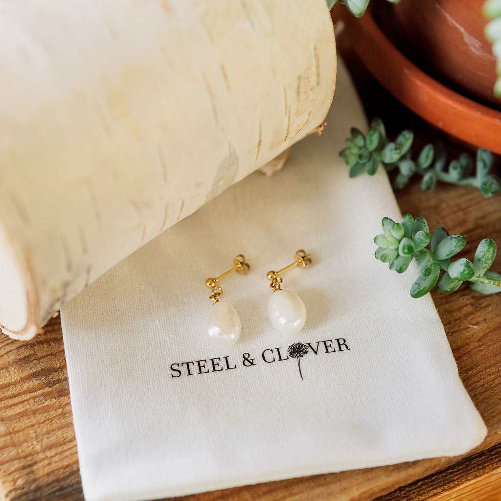 Pearl earrings on a 'Steel & Clover' branded pouch with a wooden background and plant.