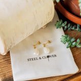 Pearl earrings on a 'Steel & Clover' branded pouch with a wooden background and plant.