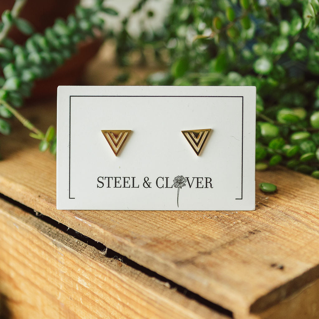 A pair of gold-plated, geometric triangle stud earrings on a presentation card that reads 'STEEL & CLOVER'.