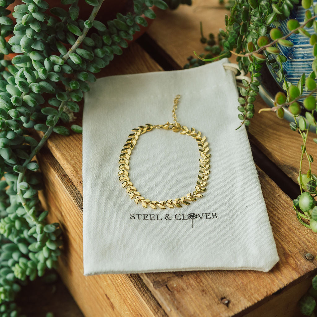 A gold-colored bracelet displayed on a beige fabric with the brand name 'Steel & Clover' embroidered on it, surrounded by green plants.