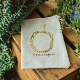 A gold-colored bracelet displayed on a beige fabric with the brand name 'Steel & Clover' embroidered on it, surrounded by green plants.
