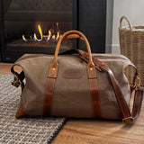 Brown travel bag with leather straps on a wooden floor in front of a fireplace.