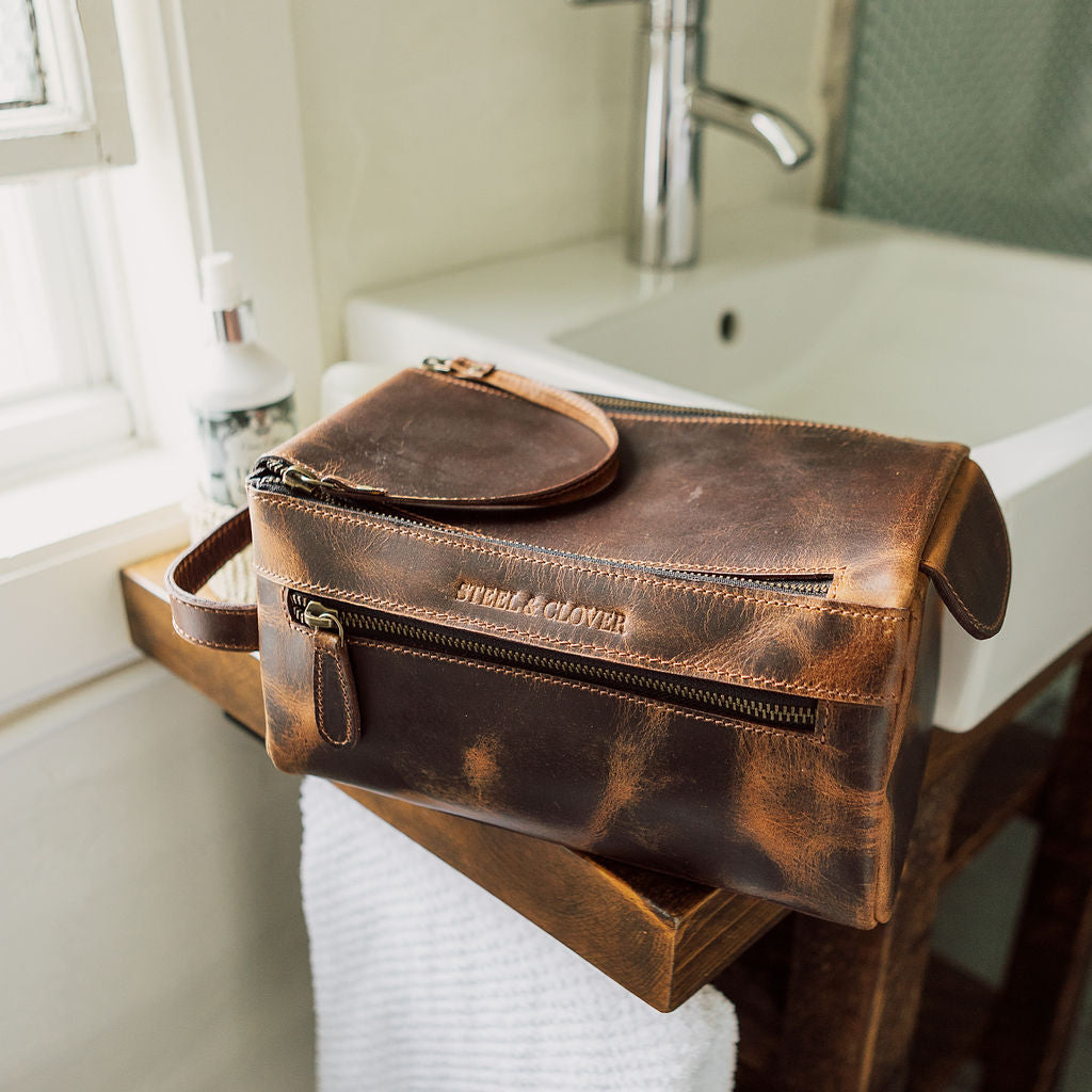 Brown leather toiletry bag on a bathroom counter with sink and window in the background