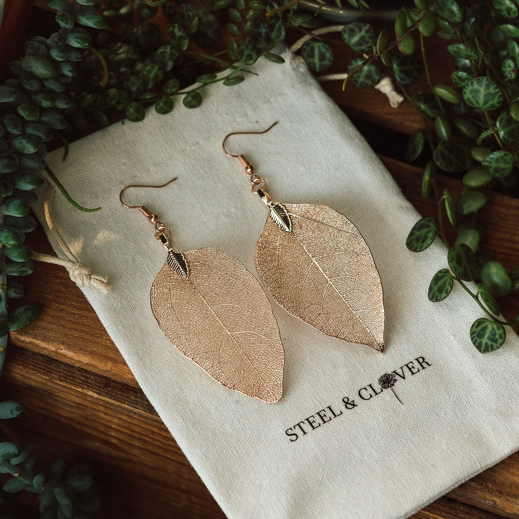 A pair of rose gold leaf-shaped earrings laid on a fabric pouch with the brand name 'Steel & Clover' embroidered on it, surrounded by green leafy foliage.