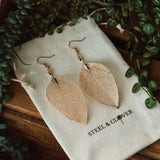 A pair of rose gold leaf-shaped earrings laid on a fabric pouch with the brand name 'Steel & Clover' embroidered on it, surrounded by green leafy foliage.