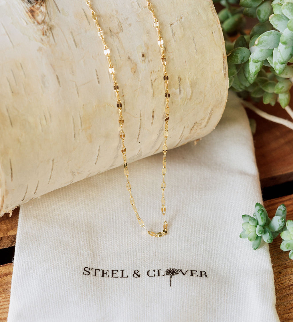 A gold-colored mirror chain necklace placed on a white textured surface with a branded cloth underneath, Steel & Clover.