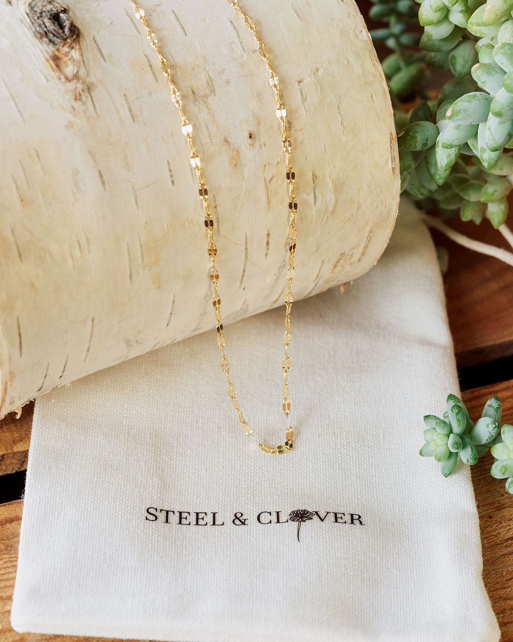 A gold-colored mirror chain necklace placed on a white textured surface with a branded cloth underneath, Steel & Clover.