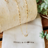 A gold-colored mirror chain necklace placed on a white textured surface with a branded cloth underneath, Steel & Clover.