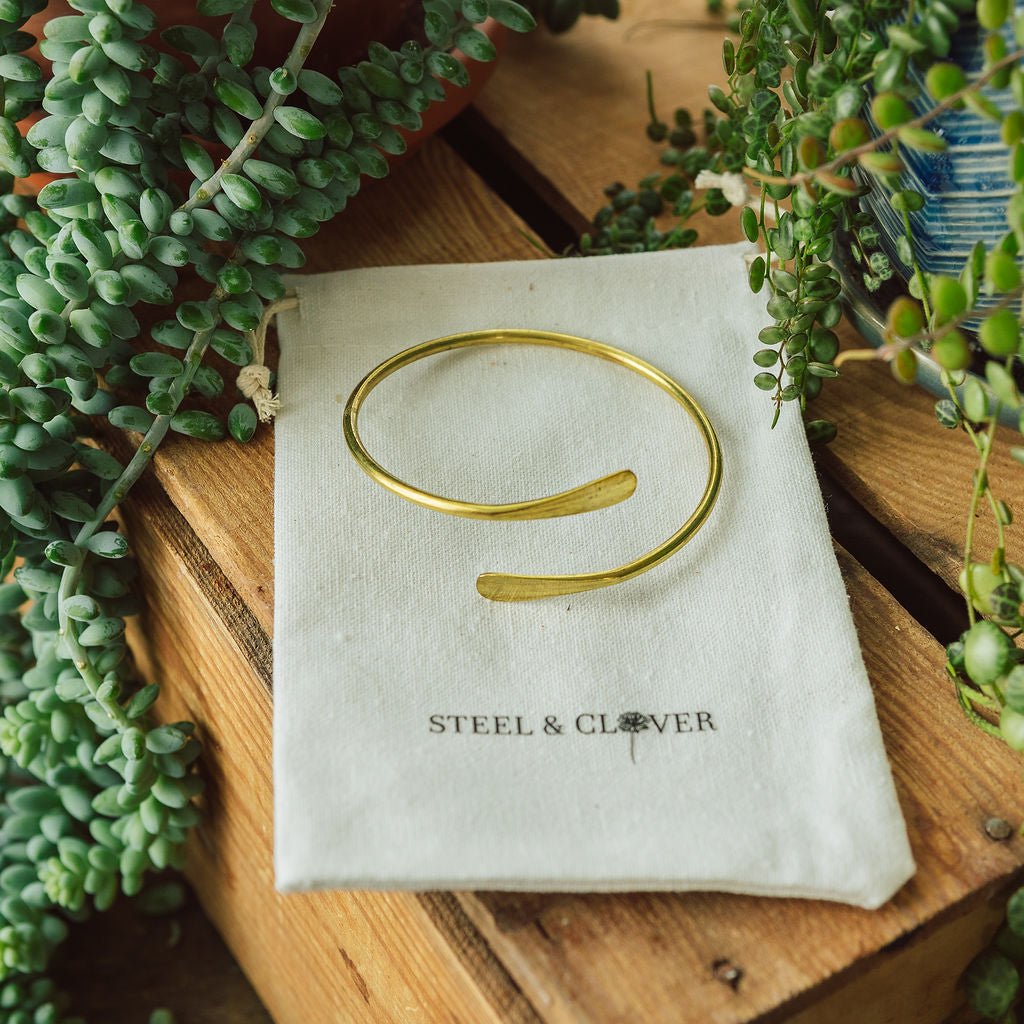 A gold-colored hammered brass bracelet on a white card with the brand name 'Steel & Clover' surrounded by greenery.