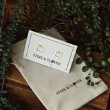 A pair of square gold-plated stud earrings on a white card with the brand name 'Steel & Clover', displayed on a wooden surface surrounded by greenery.