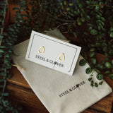 Teardrop stud earrings with a gold plating, displayed on a card with the brand name 'Steel & Clover', placed on a fabric pouch with the same brand logo, surrounded by greenery.