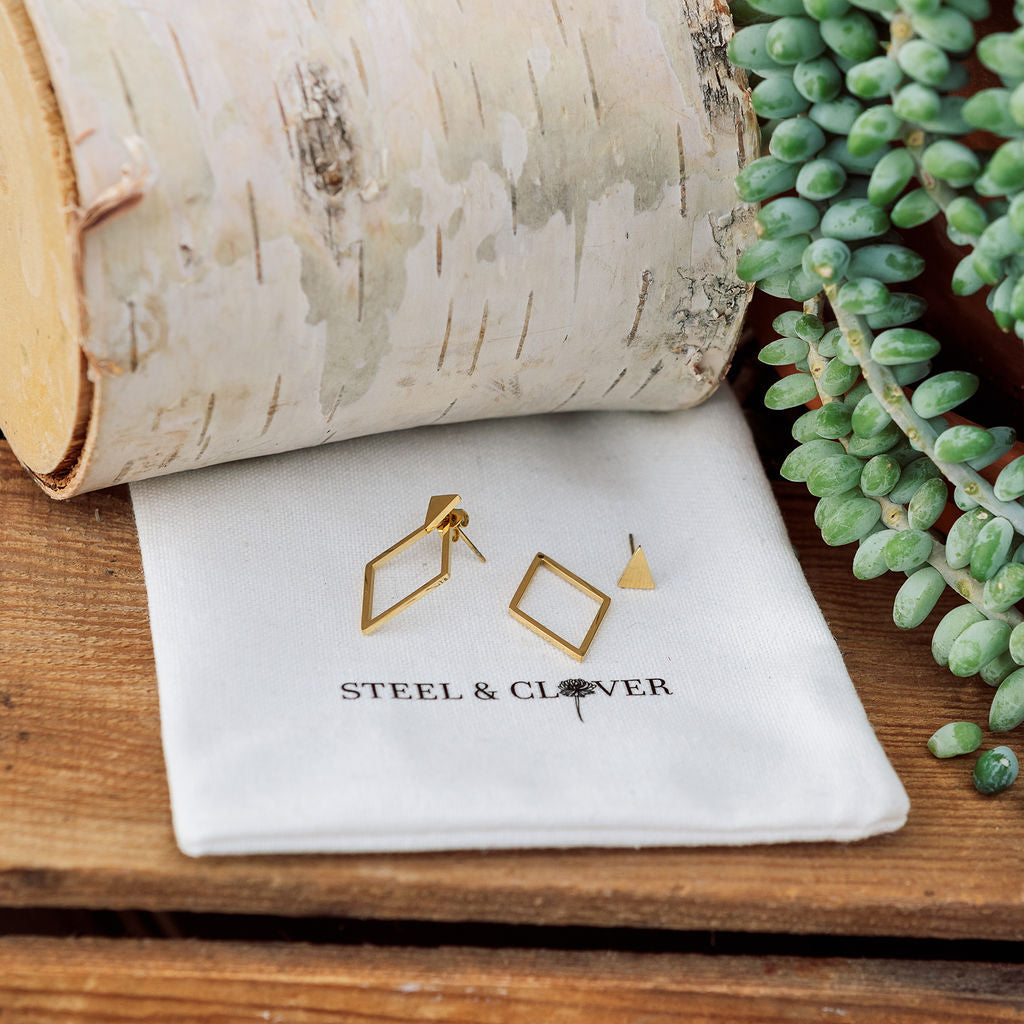 Gold geometric earrings on a white cloth with 'Steel & Clover' branding, next to a birch log and succulent plant.
