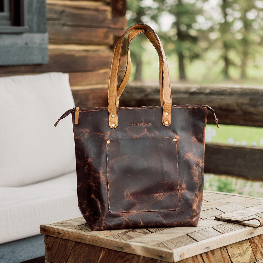 Brown leather tote bag with tan handles on a wooden surface outdoors