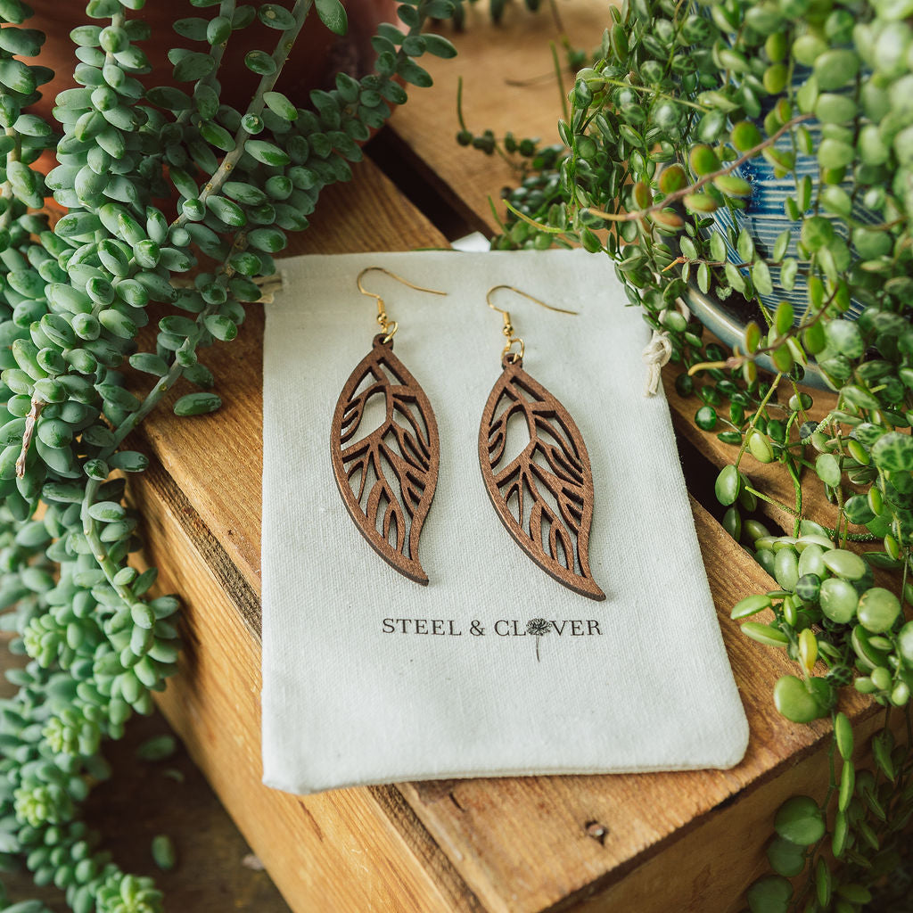 A pair of wooden leaf-shaped earrings displayed on a white fabric with the brand name 'Steel & Clover' visible, set against a backdrop of green plants.