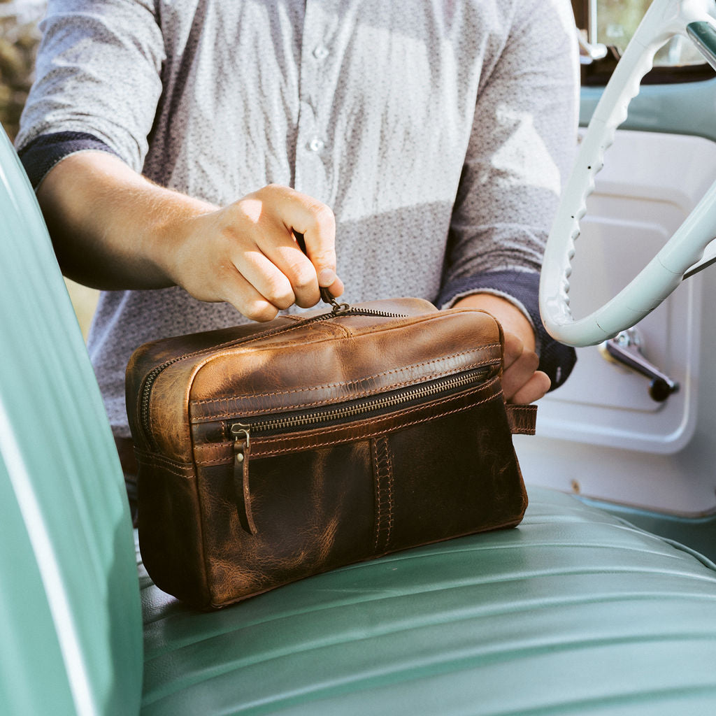 A person sitting in a vehicle's back seat, holding a brown leather dopp kit with brass zippers.