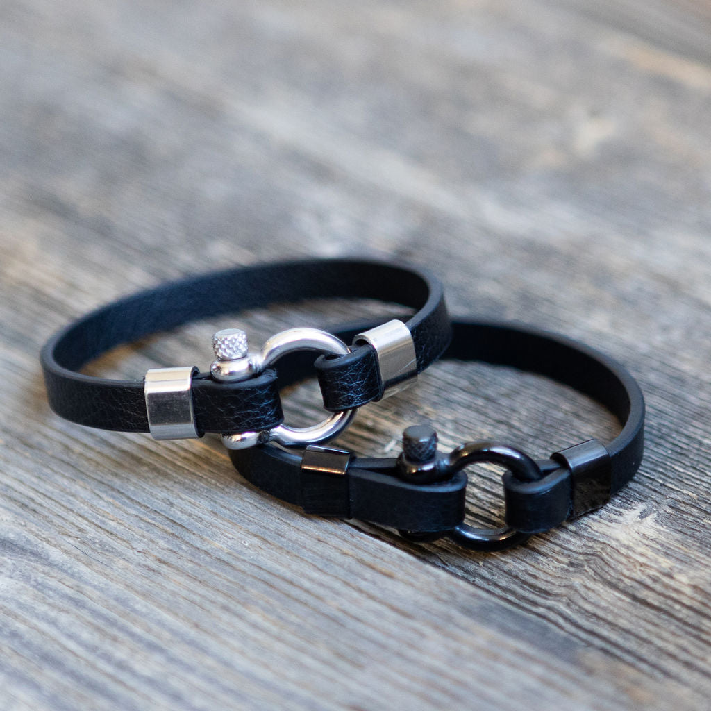 Two black leather bracelets with metal U-ring links and screw fasteners, placed on a wooden surface.