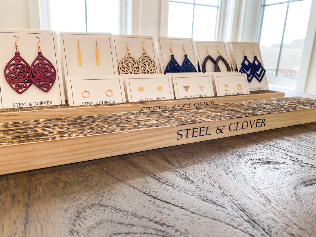 A wooden earring display stand with multiple slots, holding a variety of earrings, with the brand name 'Steel & Clover' visible on the front.