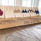 A wooden earring display stand with multiple slots, holding a variety of earrings, with the brand name 'Steel & Clover' visible on the front.